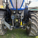 Front view of a blue agricultural tractor with large treaded tires, headlights, and front hitch on grass under a clear sky.