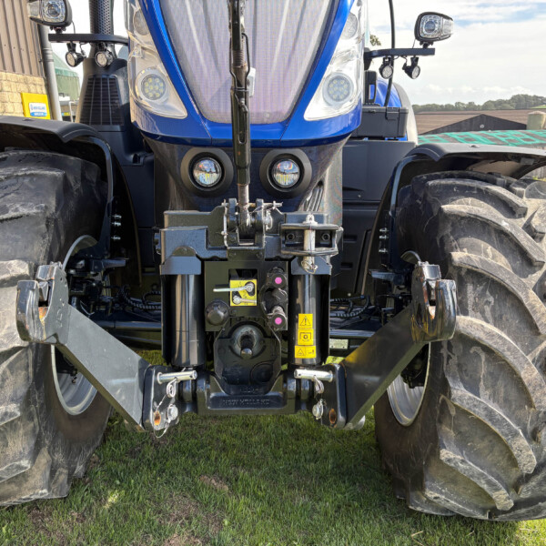 Front view of a blue agricultural tractor with large treaded tires, headlights, and front hitch on grass under a clear sky.