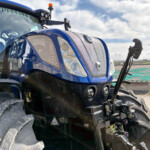 Close-up of a blue tractor front end with large treaded tires, chrome grille, and windshield showing 'RUSSELLS' against a cloudy sky