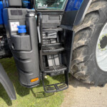 Close-up of a blue truck cab with black fuel tank, stairs, and large treaded front wheel with muddy tire and wheel bolts visible
