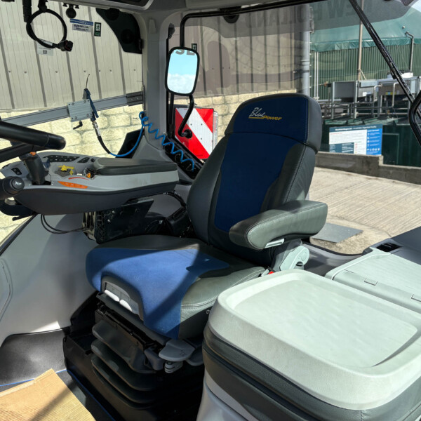 Interior view of a heavy vehicle cab with a blue driver's seat, control console, and large side mirror; exterior industrial area visible through the window.