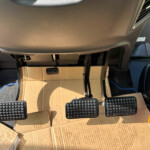 Driver’s footwell with three pedals (clutch, brake, accelerator) and exposed wiring; cardboard box resting on the floor under the dash