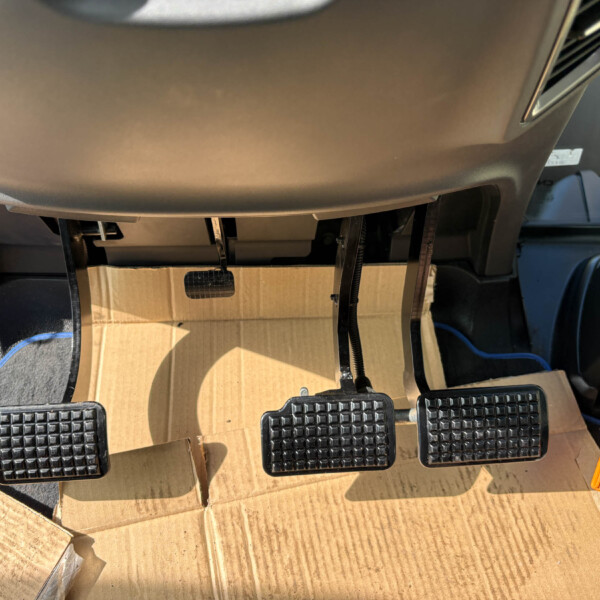 Driver’s footwell with three pedals (clutch, brake, accelerator) and exposed wiring; cardboard box resting on the floor under the dash