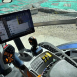 Interior of a heavy machinery cab showing a mounted tablet with control software and a joystick nearby; exterior gravel visible through the side window.