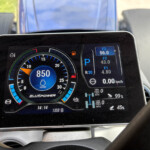 Close-up of a car's digital instrument cluster: a circular gauge with blue accents, a central blue display, and small readouts for gear (P), fuel, and other indicators.