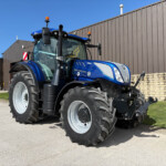 Blue New Holland tractor parked outside a brown metal industrial building on a sunny day, front view of cab and oversized tires.