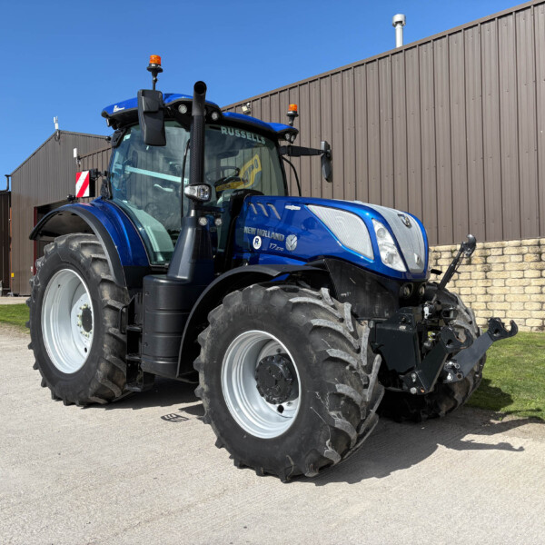 Blue New Holland tractor parked outside a brown metal industrial building on a sunny day, front view of cab and oversized tires.
