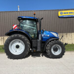 Blue New Holland tractor with large front tires parked in front of a brown corrugated metal wall, with a New Holland Agriculture sign to the right.