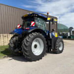 Blue agricultural tractor with oversized tires parked beside a brown metal building under a blue sky.
