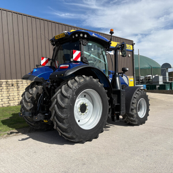 Blue agricultural tractor with oversized tires parked beside a brown metal building under a blue sky.