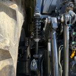 Close-up of a heavy vehicle's suspension and hydraulic system, showing a coil spring, metal linkages, and hoses between the tire and chassis.