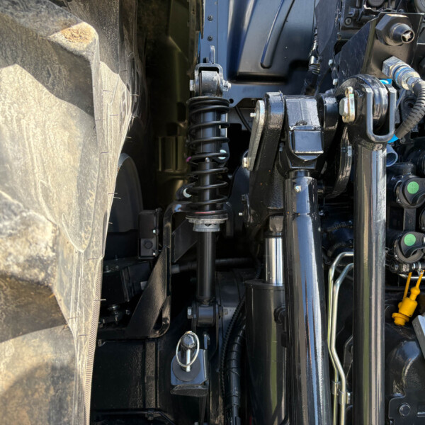 Close-up of a heavy vehicle's suspension and hydraulic system, showing a coil spring, metal linkages, and hoses between the tire and chassis.