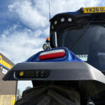 Blue tractor cab front with a yellow license plate YX26 UJS and multiple lights above the windshield near a New Holland Agriculture sign boundary.