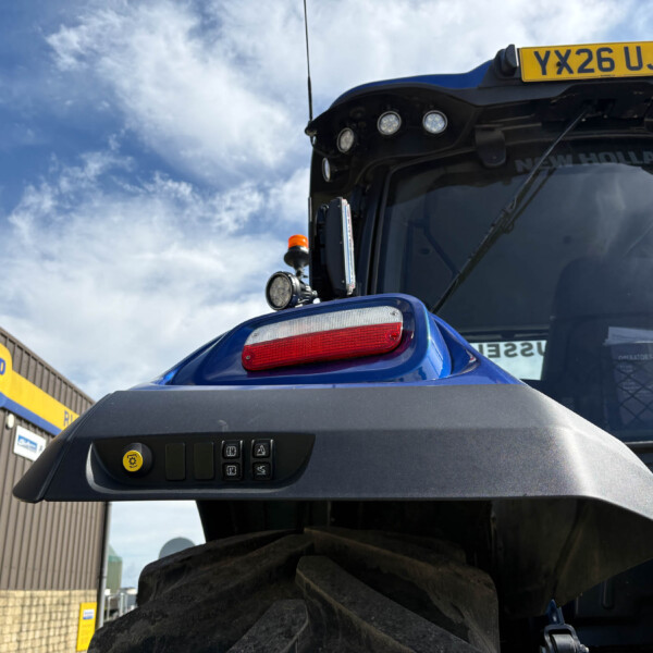 Blue tractor cab front with a yellow license plate YX26 UJS and multiple lights above the windshield near a New Holland Agriculture sign boundary.