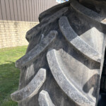 Close-up of a heavy-duty excavator/bucket with sharp, jagged teeth, dirty metallic surface, against a stone wall and grassy area background.
