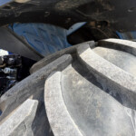 Close-up of a large dirty tire and undercarriage of heavy equipment, with a blue metal frame and exposed hydraulic components visible behind the wheel, and dirt on surfaces.
