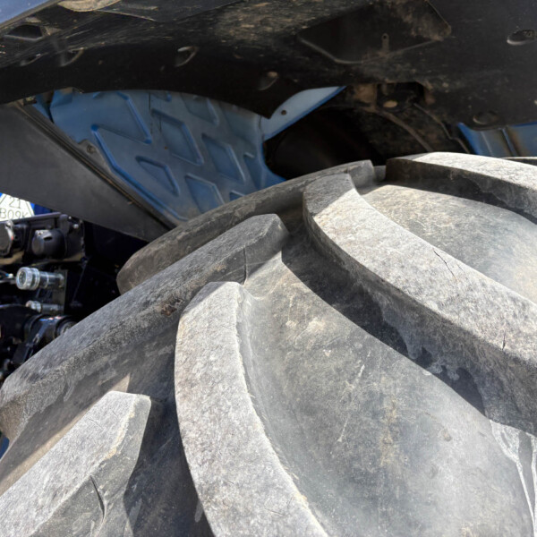 Close-up of a large dirty tire and undercarriage of heavy equipment, with a blue metal frame and exposed hydraulic components visible behind the wheel, and dirt on surfaces.