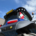 Close-up of a blue truck cab with a yellow license plate reading 'Y-X26 UJS', multiple roof-mounted spotlights, hazard striped panels, and a blue sky background.