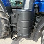 Close-up of a blue tractor cab with black step ladder and large front tire visible on the left, dirt on tire tread.