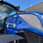 Close-up of a blue New Holland tractor cab and front grille, with an orange warning beacon on top and the word 'RUSSELLS' on the windshield against a clear blue sky.