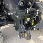 Close-up of a heavy tractor hitch with hydraulic linkages, wiring, and safety decals beside a large treaded wheel.