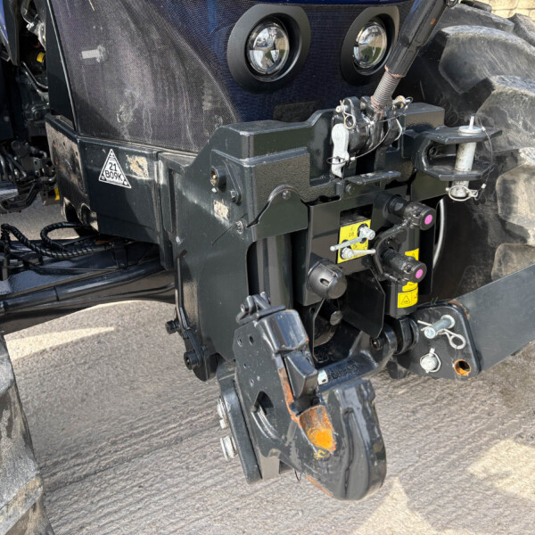 Close-up of a heavy tractor hitch with hydraulic linkages, wiring, and safety decals beside a large treaded wheel.