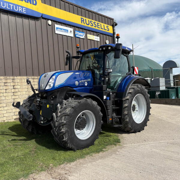Blue New Holland tractor with oversized tires parked beside a dark brown industrial building with yellow-and-blue Russell signage.