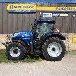 Blue New Holland tractor with oversized tires parked in front of a brown corrugated-metal building with a yellow and blue New Holland Agriculture sign and smaller logos above.