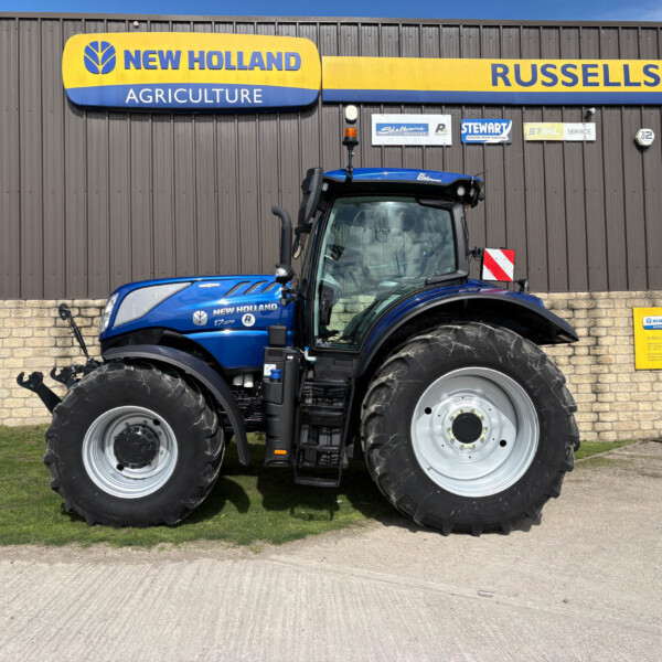 Blue New Holland tractor with oversized tires parked in front of a brown corrugated-metal building with a yellow and blue New Holland Agriculture sign and smaller logos above.