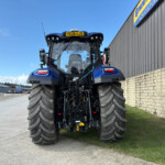 Front view of a blue agricultural tractor with oversized tires parked outdoors beside a corrugated metal building under a clear blue sky.