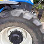 Close-up of a large tractor tire and white wheel rim, with a blue tractor body and hitch visible in the background.