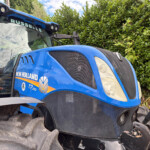 Blue New Holland T7.210 tractor front view with large tires and grill, beside a green hedge