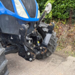 Front view of a blue tractor showing its hitch and hydraulic arms, with large black tires and a dirt-covered lower body, parked on a paved surface and green shrubbery in the background.