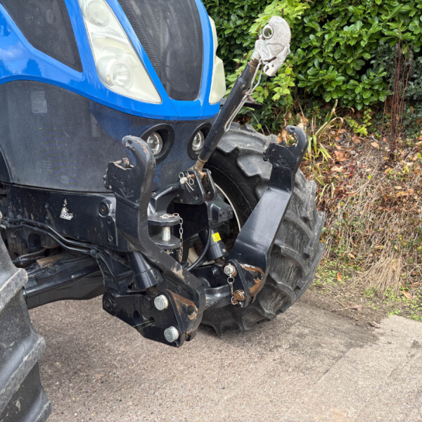Front view of a blue tractor showing its hitch and hydraulic arms, with large black tires and a dirt-covered lower body, parked on a paved surface and green shrubbery in the background.