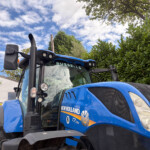 Blue New Holland tractor at a dealership, front cab and exhaust stack visible with 'RUSSELLS' on the windshield, blue sky and green hedge in the background.
