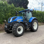 Blue New Holland tractor parked beside a tall hedge on a street, large off-road tires and orange beacon visible on top of cab.