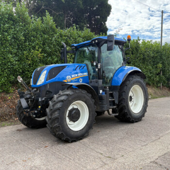 Blue New Holland tractor parked beside a tall hedge on a street, large off-road tires and orange beacon visible on top of cab.