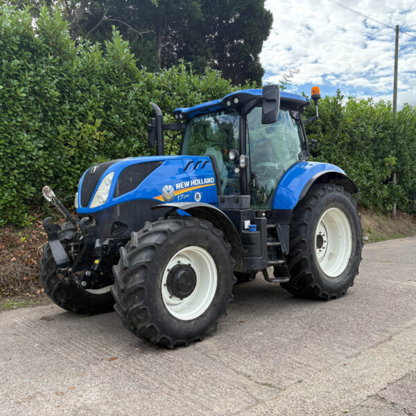 Blue New Holland tractor parked beside a tall hedge on a street, large off-road tires and orange beacon visible on top of cab.