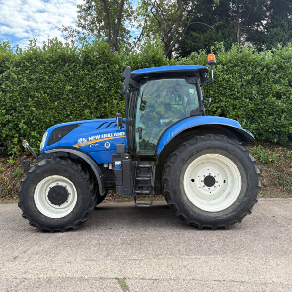 Blue New Holland tractor parked along a tall green hedge, front and cab visible with large off-road tires and an orange beacon on top of the cab.