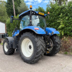 Blue agricultural tractor parked outdoors in front of a building, showing its large front tires and orange warning beacon on top.