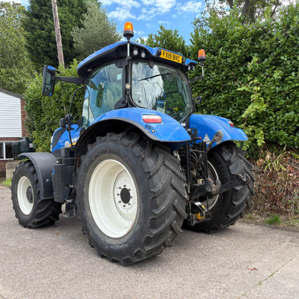 Blue agricultural tractor parked outdoors in front of a building, showing its large front tires and orange warning beacon on top.