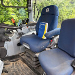 Blue tractor cab seat with operator's manual in pocket and control levers nearby, greenery visible through window behind the seat