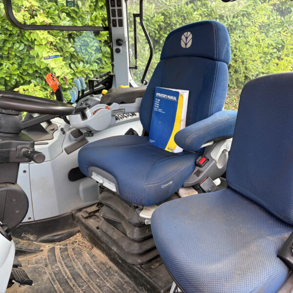 Blue tractor cab seat with operator's manual in pocket and control levers nearby, greenery visible through window behind the seat
