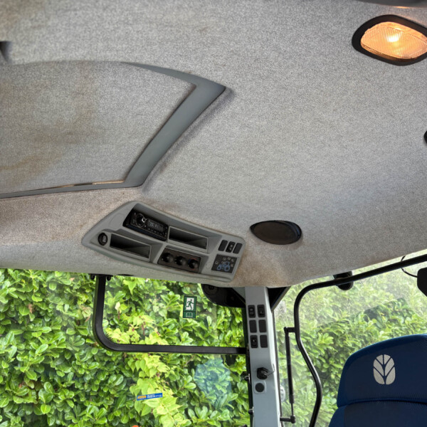 Interior of a vehicle cab (tractor/utility truck): gray fabric ceiling, overhead control panel, sun visor, rearview mirror, and round speaker; green hedge visible through the window and a blue seat with a leaf logo in the foreground.