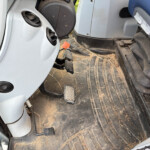 Driver's footwell with accelerator, brake and clutch pedals, dusty rubber mat with straw and dirt on the floor.