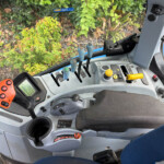 Inside tractor cab: blue gear levers, yellow controls, dashboard display, view of plants outside the window