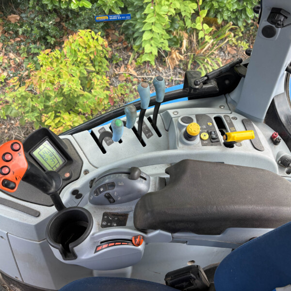 Inside tractor cab: blue gear levers, yellow controls, dashboard display, view of plants outside the window