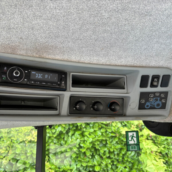 Dashboard of a vehicle cab showing a radio head unit, climate controls, and an open storage slot; outside window shows green foliage and an emergency exit sign.