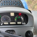 Dashboard view of a vehicle showing blue function buttons along the top, a steering wheel, and analog gauges; digital readout shows 4792.9 miles (or hours).
