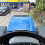 View from inside a blue tractor cab looking toward a New Holland Russell's Agriculture shop with a black car parked in front.